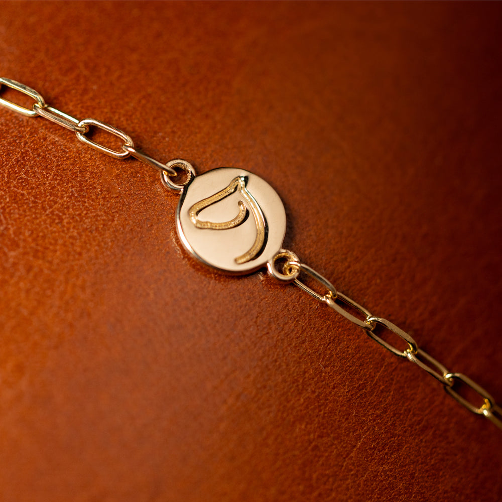 A close-up image of a gold bracelet with a small circular medallion charm attached to a paper clip style chain with a lobster clasp, displayed on a brown background.
