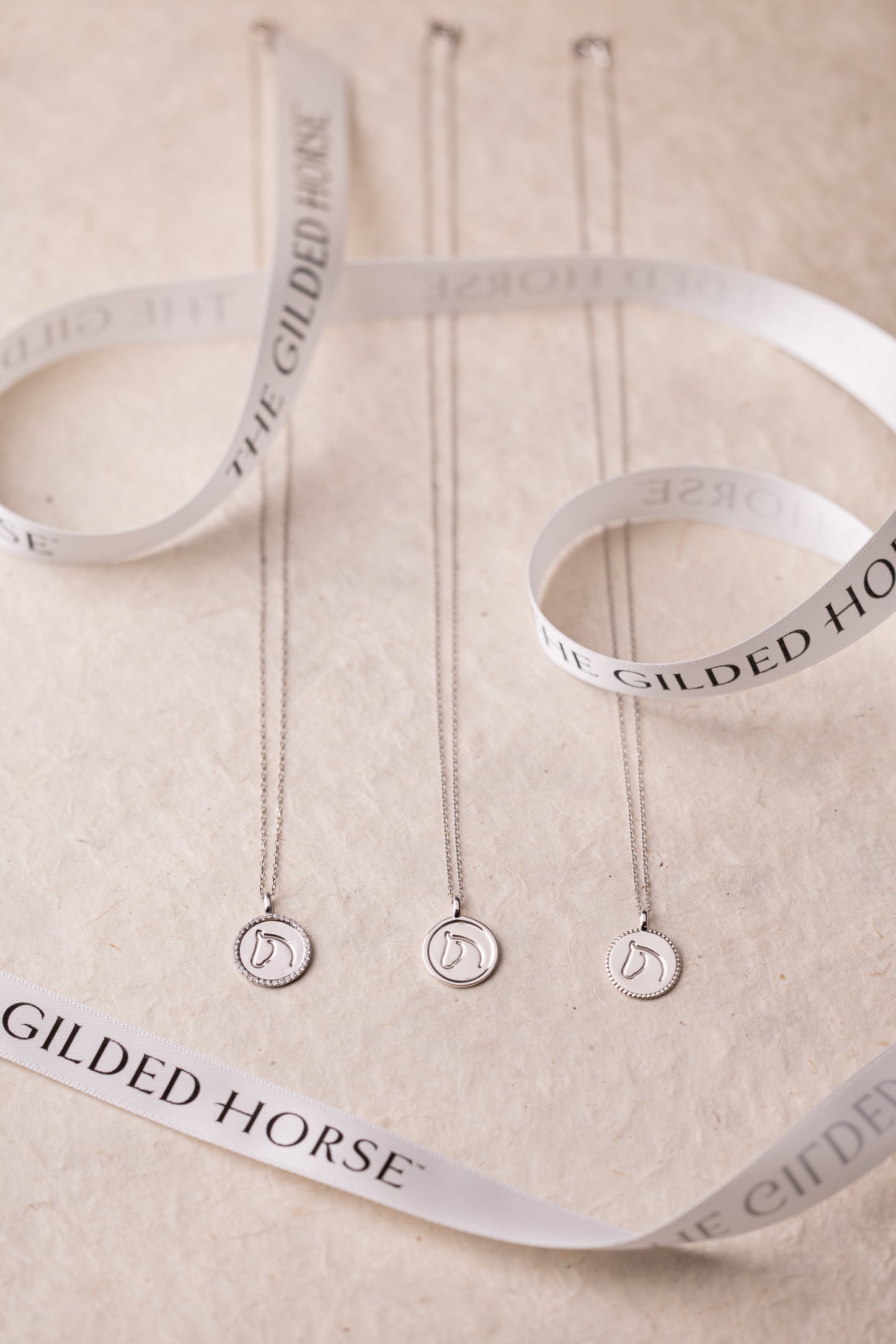 Three necklaces with circular pendants on white ribbons with 'Gilded Horse' branding.