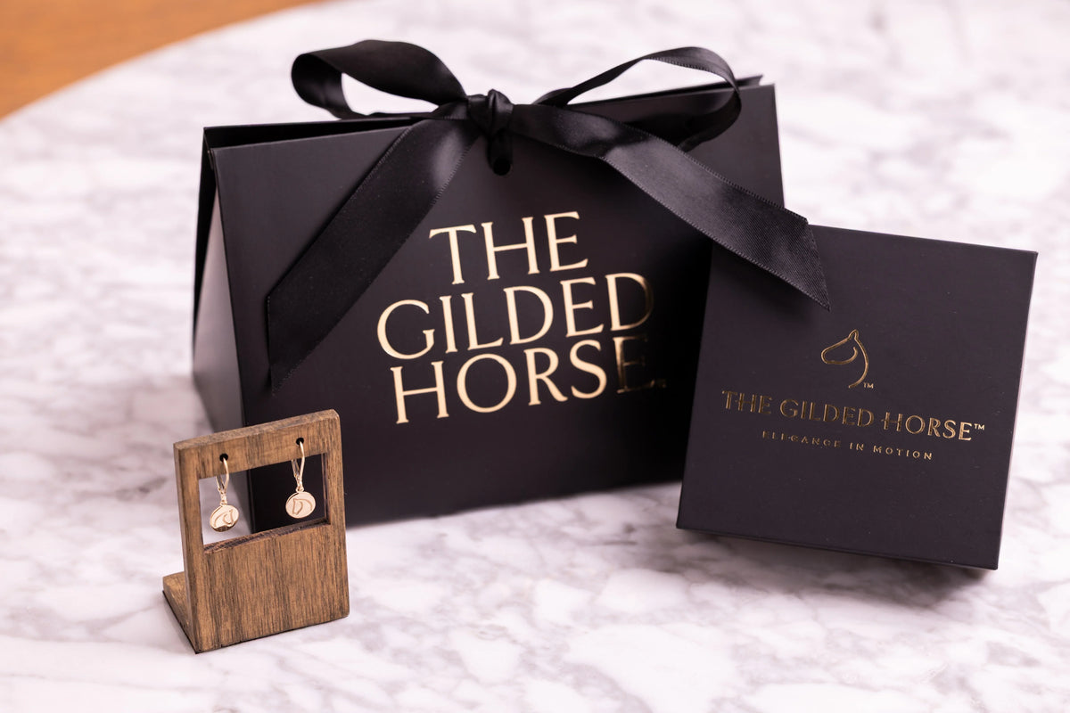 Christmas gifts for equestrians from The Gilded Horse- gold Medallion dangle earrings beside a black ribbon-tied gift bag and logo jewelry box