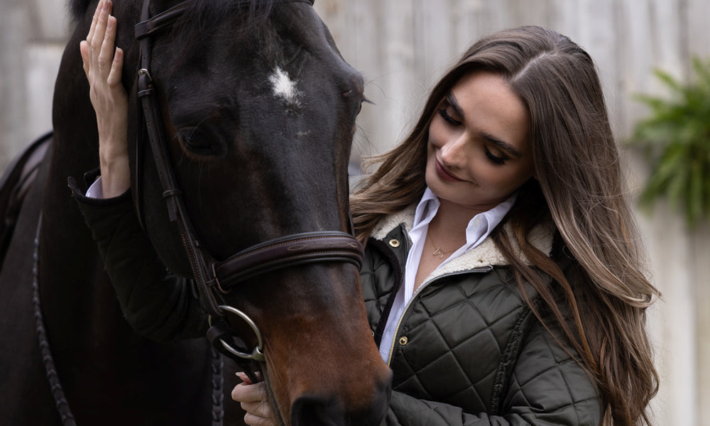 The Gilded Horse: woman in a quilted jacket and horse-head necklace cradling a dark bay horse
