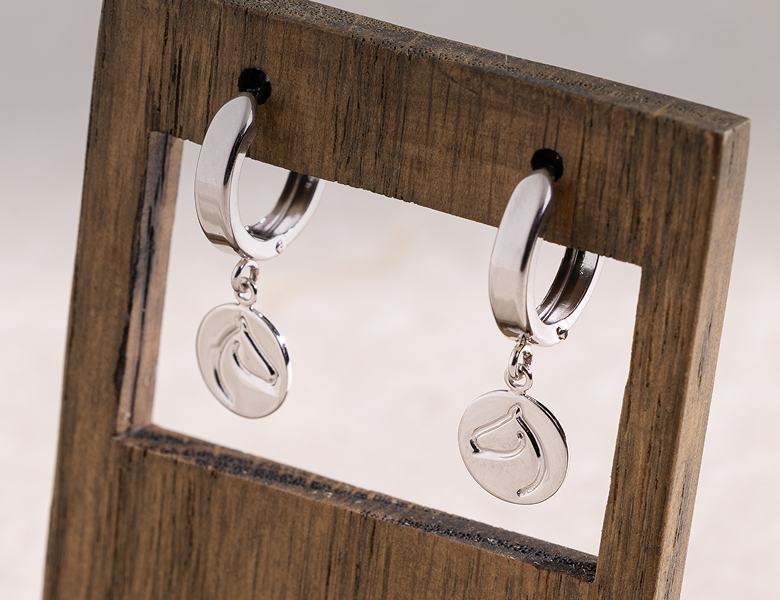 Silver-tone huggie hoop earrings with engraved horse-head medallion drops on a wood stand