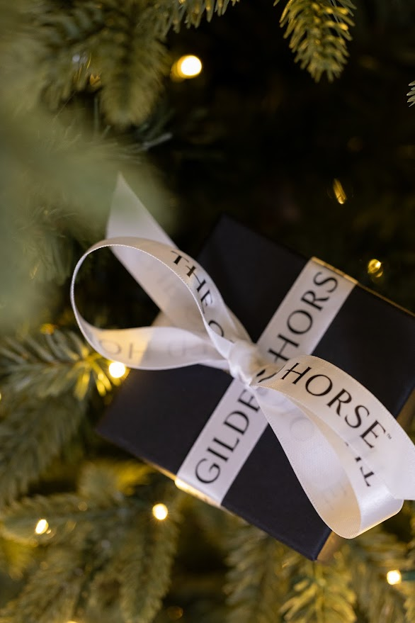 Black The Gilded Horse gift box tied with a branded white ribbon on a lit holiday tree