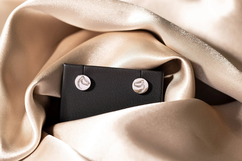Gold horse-head circle medallion stud earrings on a black stand atop champagne silk