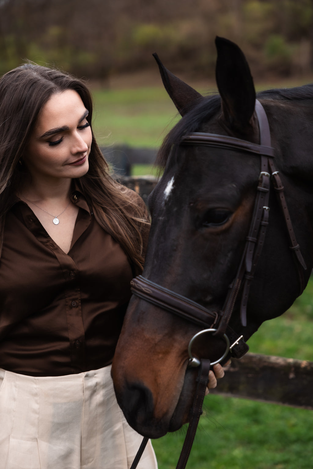 Woman in a brown shirt and beige pants, with a necklace from The Gilded Horse, standing next to a horse in an outdoor setting.