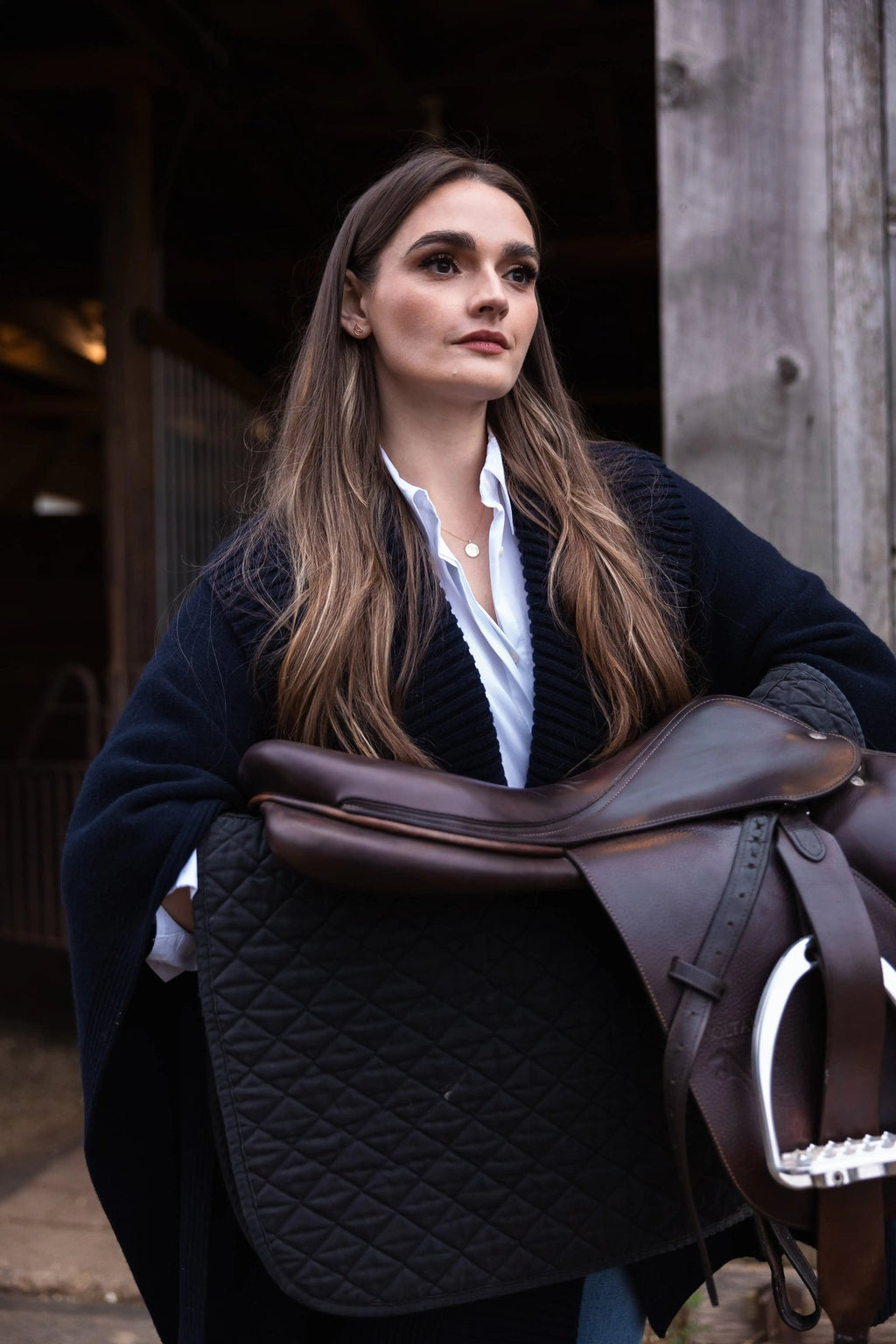 Woman in a barn holding a leather saddle, wearing The Gilded Horse jewelry