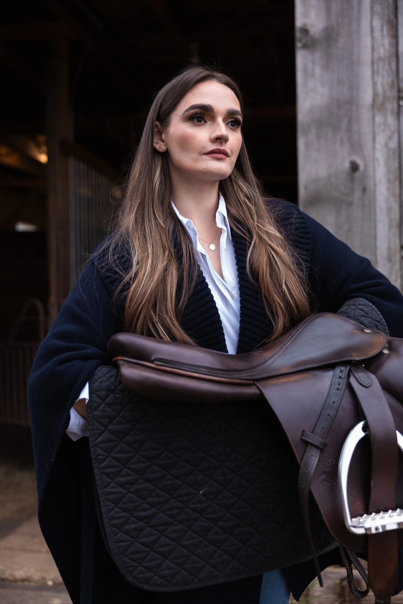 Woman in a barn holding a leather saddle, wearing The Gilded Horse jewelry