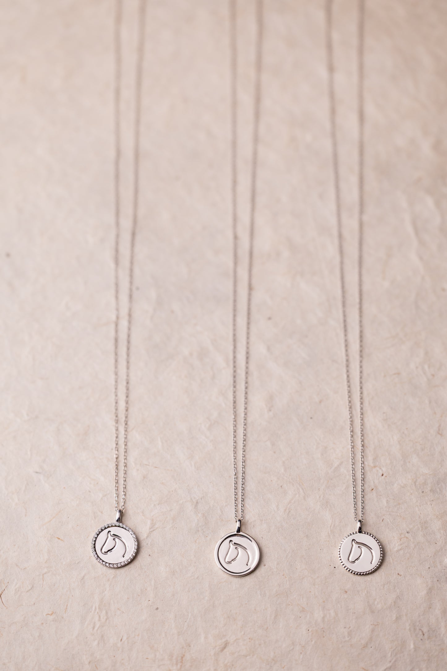 Three silver necklaces with circular horse pendants on a beige textured background.