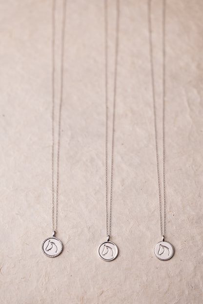 Three silver necklaces with circular horse pendants on a beige textured background.