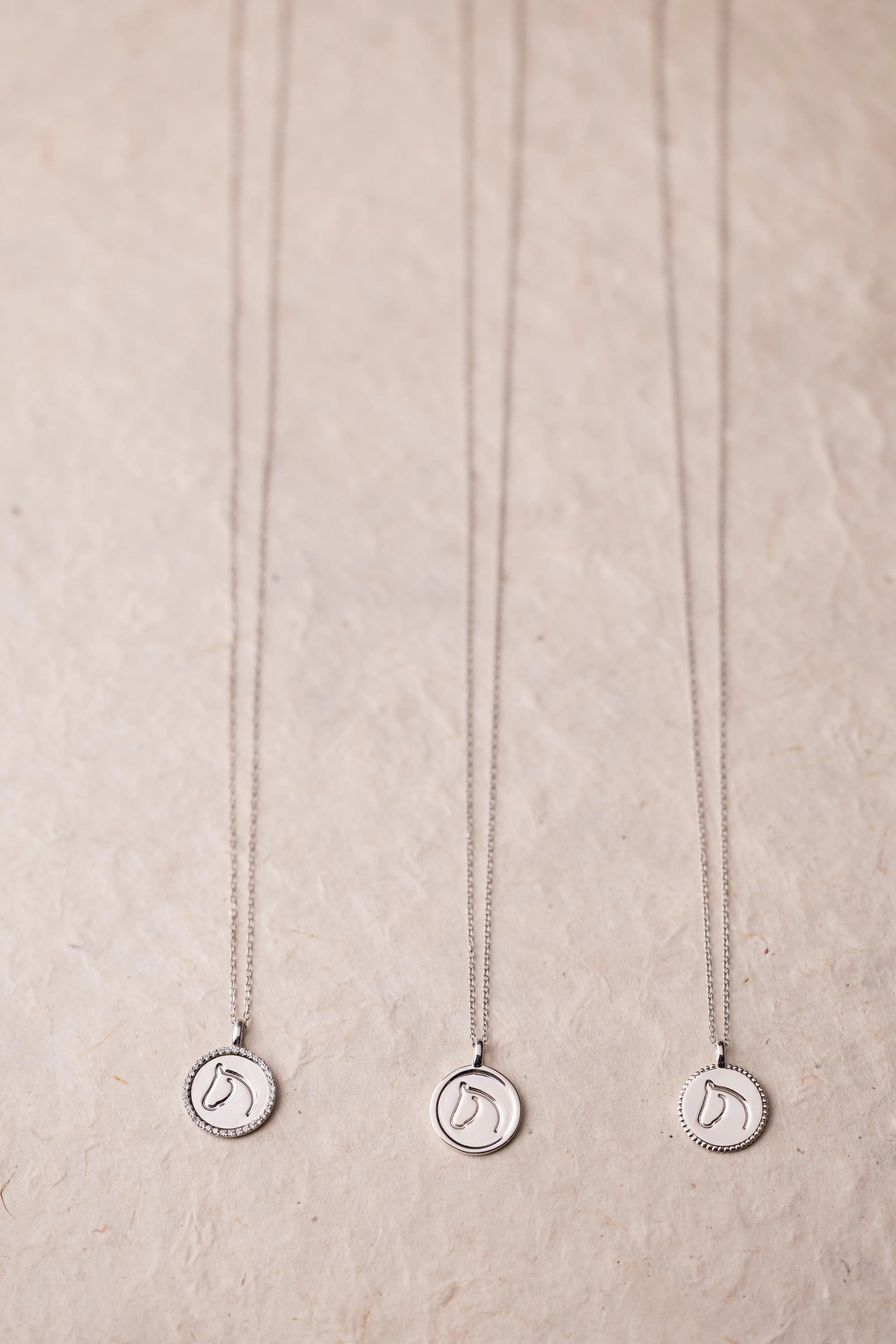 Three silver necklaces with circular horse pendants on a beige textured background.