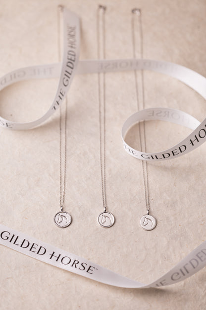 Three necklaces with circular pendants on white ribbons with 'Gilded Horse' branding.
