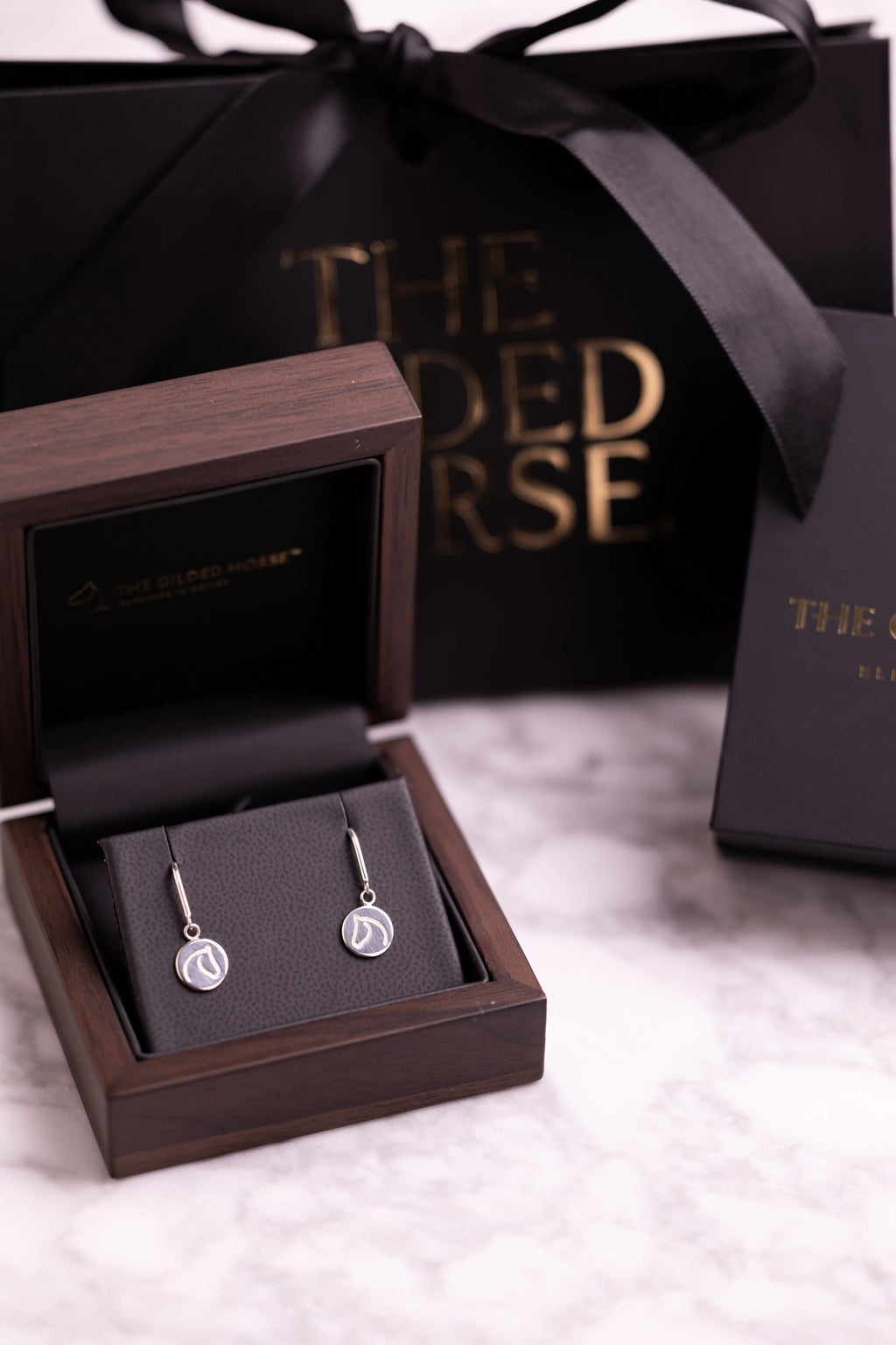 Silver-tone circle medallion earrings in a wooden box beside The Gilded Horse gift packaging