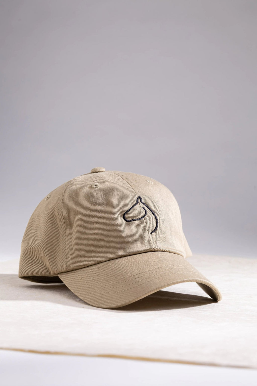 The Gilded Horse khaki cap with embroidered horse-head logo on a soft gray backdrop
