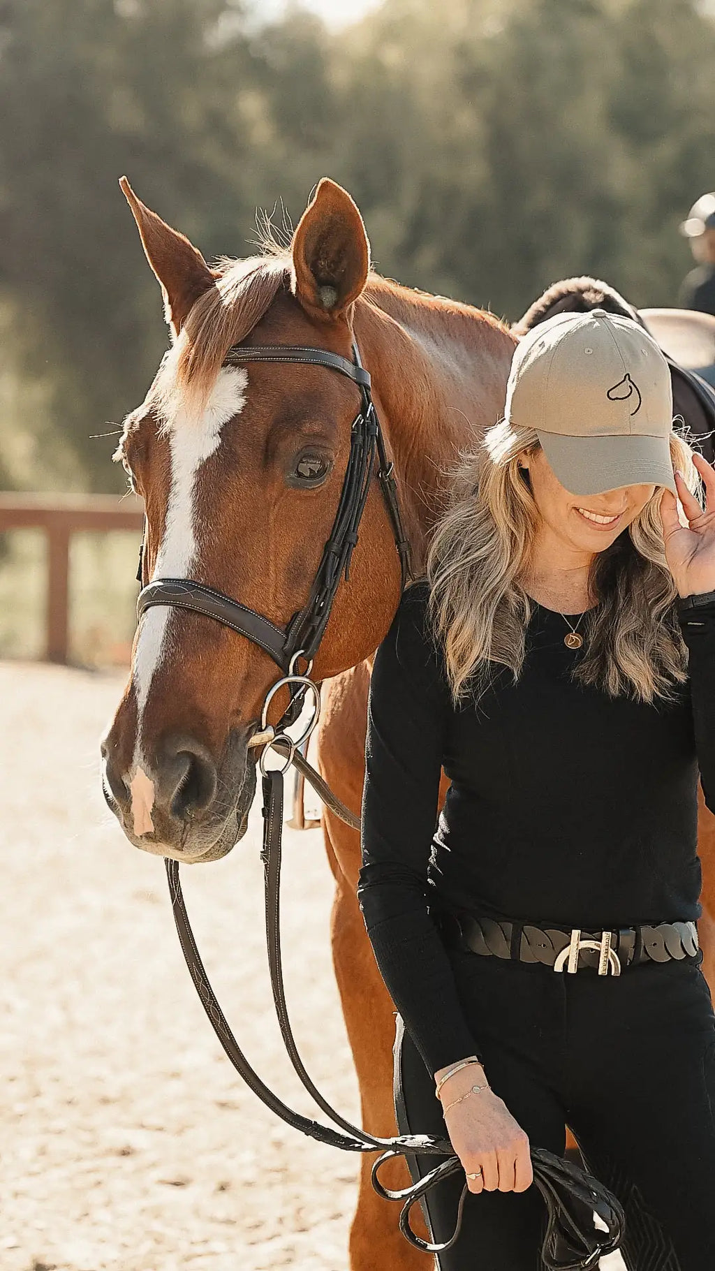 Woman in black leading a chestnut horse and wearing The Gilded Horse khaki logo cap outdoors