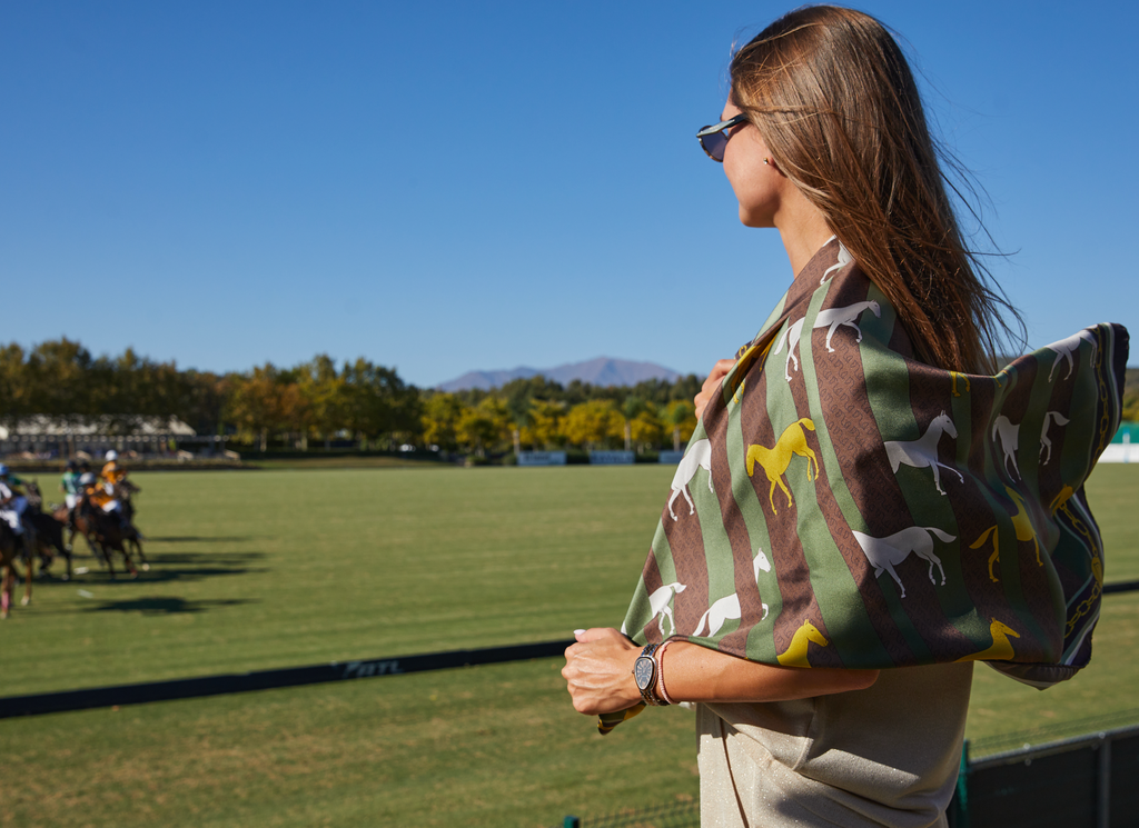 A woman wearing sunglasses stands on a horse racing track, wrapped in a green and brown striped scarf with white and yellow horse silhouettes. In the background, horse racers are visible on a green field under a clear blue sky.