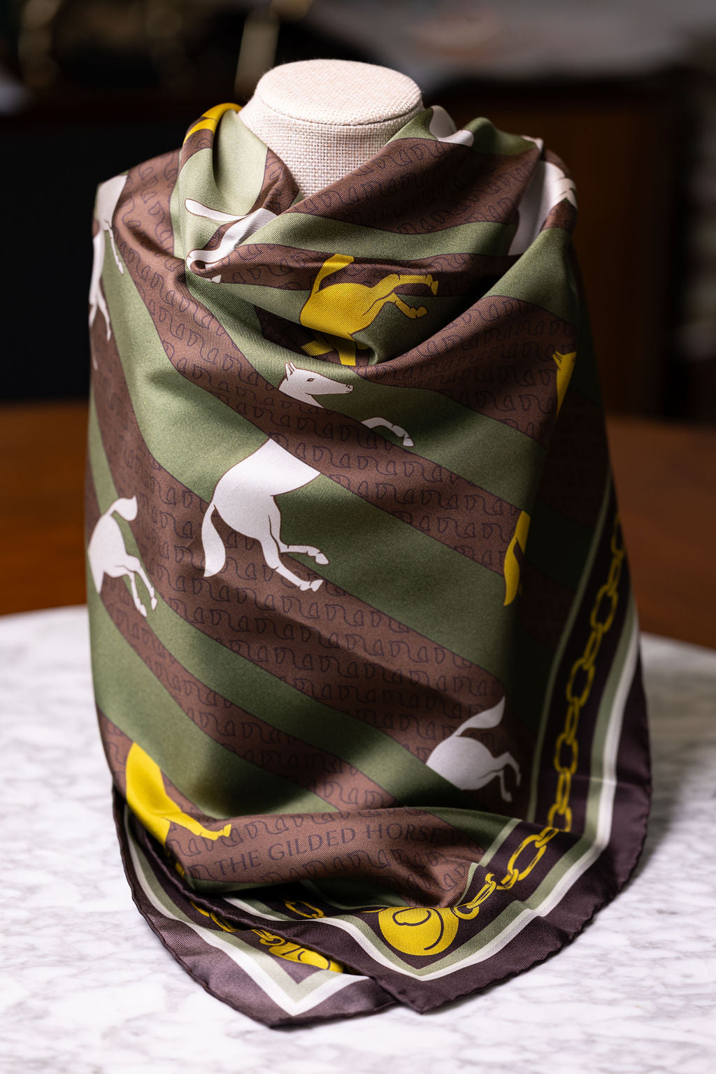 A square silk scarf draped on a mannequin torso, featuring a pattern of white horses on diagonal olive green and dark brown stripes, with a yellow border and chain motif.