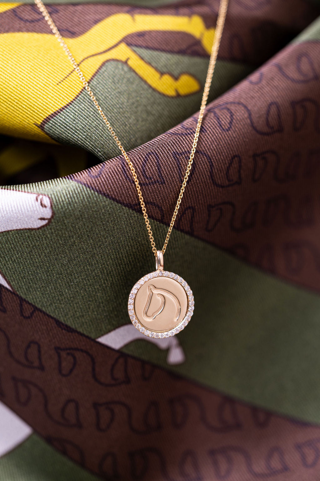 A gold pendant necklace with a circular medallion featuring a horse head design, displayed on a patterned silk scarf with green, brown, and yellow tones.
