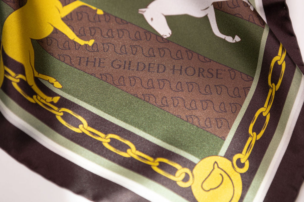 A close-up of a silk scarf featuring The Gilded Horse logo, with a brown center, olive green stripes, a yellow horse silhouette, and a gold chain border.