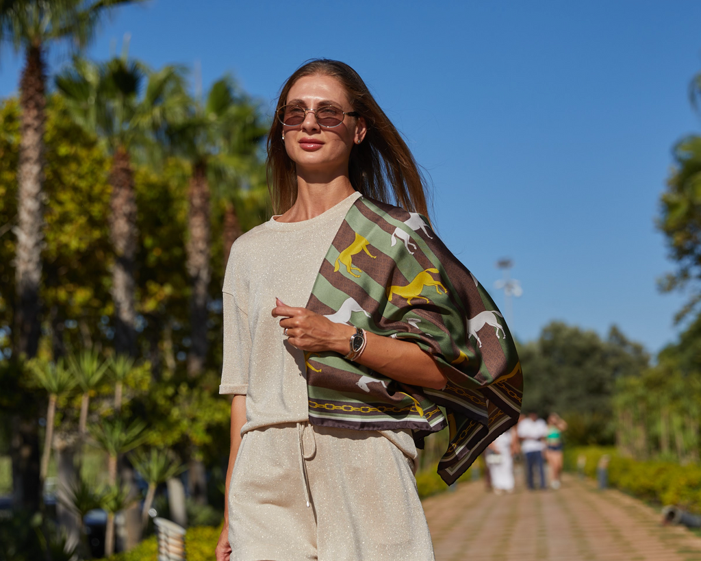 A woman walking outdoors on a sunny path wearing a beige knit outfit and carrying a green, brown, and yellow patterned scarf.